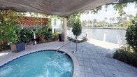 This inviting outdoor amenity space is defined by its prime waterfront setting and relaxed tropical ambiance. At the heart of the space sits a circular hot tub with active jets, framed by a distinctive concrete overhead canopy supported by elegant columns, which provides shade while maintaining an open, airy feel. The surrounding patio is finished with attractive paver tiles, offering a clean and durable surface ideal for barefoot relaxation.

Lush, mature tropical landscaping borders the space, featuring a vibrant mix of palms, flowering plants, and dense foliage that creates a sense of privacy while enhancing the resort-like atmosphere. A decorative perforated brick wall adds an architectural accent to one side of the space.

Beyond the patio, a white railed waterway boundary opens up to sweeping views of a calm canal or inlet, with palm trees and neighboring waterfront properties visible in the distance. The proximity to the water creates a naturally breezy and serene environment. This outdoor space is well-suited for leisure and relaxation, combining functional amenities with the natural beauty of a coastal Florida setting.