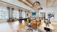 This well-appointed amenity lounge offers a sophisticated and welcoming atmosphere, thoughtfully designed for both socializing and relaxation. The open-concept layout accommodates a variety of activities across its generously sized single floor. Exposed natural wood beams traverse the high ceilings, adding warmth and architectural character to the bright, airy interior. Underfoot, wide-plank hardwood flooring transitions seamlessly into large-format tile, defining different functional zones within the space.

A full-size billiards table anchors the entertainment area, positioned beneath striking oversized bronze dome pendant lights that serve as bold focal points. Adjacent to the game area, a stylish wet bar features patterned blue-and-white tile backsplash, wood cabinetry, bar seating, and a wall-mounted television — ideal for casual gatherings. Multiple curated seating arrangements include a leather sofa, tufted armchairs, and velvet lounge chairs, creating intimate conversation areas throughout.

Expansive floor-to-ceiling windows with sheer drapery and wood-slat blinds line the perimeter, flooding the space with natural light and offering open views of the surrounding streetscape. The overall aesthetic blends contemporary design with warm, natural textures, resulting in an inviting and refined communal environment.
