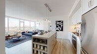 This thoughtfully designed single-floor residence showcases a contemporary open-concept layout bathed in natural light. The expansive living and dining area is anchored by floor-to-ceiling windows that frame sweeping city views, creating a bright and airy atmosphere throughout. Light hardwood flooring runs seamlessly across the main living spaces, complementing the clean white walls and modern finishes.

The kitchen is a standout feature, offering white shaker cabinetry, a herringbone tile backsplash, stainless steel appliances, and a large center island with quartz countertops — ideal for both cooking and casual dining. Elegant pendant lighting adds a refined touch overhead. The dining area is adjacent, featuring its own statement chandelier and continued access to the panoramic windows.

The primary bedroom is generously sized, offering a walk-in closet and a serene, neutral palette. A second bedroom also includes a walk-in closet, providing ample storage. Two bathrooms serve the residence: one features a glass-enclosed walk-in shower with a floating vanity and clean tile work, while the second includes a soaking tub with herringbone wall tiling. Together, the rooms offer a cohesive blend of modern design and functional comfort throughout.