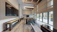 This contemporary amenity lounge is designed to impress, combining sophisticated architectural details with a versatile, social atmosphere. The space is anchored by dramatic double-height ceilings adorned with a distinctive slatted wood ceiling feature and recessed lighting, lending warmth and visual interest overhead. Floor-to-ceiling windows and large sliding glass doors flood the interior with abundant natural light while offering direct access to an outdoor courtyard, seamlessly blending indoor and outdoor living.

The lounge is thoughtfully organized into distinct activity zones. A central games area is equipped with a full-size pool table and a shuffleboard table, catering to competitive and casual play alike. An arcade console coffee table adds a playful touch to the seating area, which features plush neutral sofas accented with teal throw pillows, arranged around a sculptural concrete coffee table. A sculptural multi-bulb chandelier serves as an artistic focal point above this lounge zone.

A long communal high-top table with bar stools provides a more social or co-working setting, while a dedicated workstation counter with seating lines one wall, accompanied by artistic pendant lighting. A mosaic tile accent wall and a wall-mounted flat-screen TV complete the entertainment-focused lounge area. The refined, neutral palette of warm taupes, greys, and natural wood tones creates a cohesive and inviting ambiance throughout.