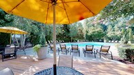 Located at 10311 Falls Mill Dr in Raleigh, NC, this outdoor amenity space centers around a generously sized swimming pool with crystal-clear blue water, framed by an expansive concrete sun deck lined with rows of lounge chairs. The pool area is enveloped by mature trees and lush greenery, creating a private, resort-like atmosphere that provides natural shade and a tranquil backdrop throughout the day.

Adjacent to the pool, a substantial wood deck serves as a social hub, featuring a white pergola structure that offers partial shade over multiple seating arrangements. Cast-iron tables paired with woven chairs are shaded by bold yellow umbrellas, creating a vibrant and inviting outdoor dining environment. The deck is also equipped with grilling stations, making it well-suited for outdoor entertaining.

A single-story clubhouse building with classic architectural detailing, including a gabled roofline and neutral exterior, anchors one side of the pool area and provides functional support facilities. Ornamental iron fencing defines the perimeter of the deck, complemented by manicured landscaping with flowering crepe myrtles and carefully arranged planters. The overall ambiance is bright, open, and welcoming, ideal for warm-weather relaxation and community gatherings.