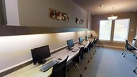 This single-floor computer lab is thoughtfully designed to provide a functional and comfortable workspace for multiple users. A long, continuous built-in desk runs along the main wall, accommodating a row of desktop computer workstations, each paired with an ergonomic rolling chair, offering ample space for individual use. Warm under-shelf lighting runs beneath a horizontal accent rail along the wall, casting a soft ambient glow across the work surface and contributing to a focused yet inviting atmosphere. The walls are finished in a neutral taupe tone, complemented by a contrasting darker horizontal band that adds a subtle architectural detail. A series of distinctive metallic wall art pieces are mounted above the workstations, introducing an artistic element that elevates the room beyond a purely utilitarian space. Natural light filters in through a window along the side wall, brightening the room and reducing the sense of enclosure. Carpeted flooring throughout adds acoustic comfort, making the space conducive to quiet, concentrated work. Overhead ventilation ensures a comfortable environment during extended use. Overall, this computer lab strikes a balance between practical functionality and polished design, making it well-suited as a shared resource or business center facility.