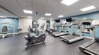 Located at 90 Gas Light Dr in Weymouth, MA, this fitness center offers a well-appointed, single-floor workout facility designed to accommodate a wide range of exercise needs. The space features a drop-panel ceiling with recessed fluorescent lighting that evenly illuminates the entire floor area, which is finished with durable rubber flooring suited for high-traffic athletic use.

The cardio zone is equipped with multiple treadmills, stationary bikes, and elliptical machines, several of which are positioned in front of wall-mounted televisions for an engaging workout experience. A dedicated strength training area houses a selection of selectorized weight machines, including a leg press and cable systems, providing options for both beginner and experienced users. The free weight section features a full rack of dumbbells alongside a adjustable weight bench, catering to a variety of strength training routines.

Large wall mirrors are strategically placed throughout the space, enhancing the sense of openness and allowing users to monitor their form during exercise. Accent walls in a cool blue tone add a modern, energetic feel to the otherwise neutral interior palette. The facility also includes built-in storage cabinetry and provides convenient access via a clearly marked exit. A glimpse of an adjacent recreation area with a billiards table is visible, suggesting additional amenity spaces nearby.