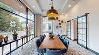 Located at 320 S Walker St in Cary, NC, this single-floor amenity space offers a thoughtfully designed environment for collaboration, focused work, and private meetings. The centerpiece is a spacious conference room anchored by a substantial live-edge wood table surrounded by tufted navy leather chairs, topped with striking oversized bronze pendant lights. Floor-to-ceiling black-framed windows flood the room with natural light and provide open views of the surrounding streetscape. Decorative metallic disc wall art and sliding barn-style glass doors add a sophisticated, contemporary touch.

Adjacent to the conference room, a dedicated co-working nook features a long communal wood countertop with blue seating, illuminated by a stylish sputnik-style brass sconce. Exposed concrete columns throughout the space provide an industrial contrast to the clean white walls and patterned carpeting. A cozy lounge corner offers a plush armchair beneath a large vintage-style world map print, ideal for quiet reading or informal conversation.

Additional private meeting rooms include a round-table space adorned with octagonal mirrors and a buffet credenza, creating an intimate yet polished setting. Black-framed steel-and-glass interior doors unify the design language across the space, while lush greenery and curated artwork complete the inviting, professional ambiance.