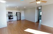 This single-floor home offers an open and airy layout that seamlessly connects the living and kitchen areas, creating a welcoming and functional space. The kitchen is outfitted with stainless steel appliances, including a range, microwave, and refrigerator, complemented by crisp white cabinetry and light stone countertops. The generous open living area flows naturally from the kitchen, making it well-suited for both everyday living and entertaining. Warm hardwood-style flooring runs throughout, adding a sense of cohesion and a touch of elegance to the interior. Crown molding lines the ceilings, lending a refined architectural detail, while recessed lighting and a classic ceiling fan with a light fixture ensure both comfort and ample illumination. Neutral gray walls provide a versatile backdrop that pairs well with a variety of décor styles. Interior doorways lead to additional rooms, hinting at a thoughtfully designed floor plan that balances open communal spaces with private quarters. Natural light filters into the space, enhancing the bright and clean aesthetic that defines this well-maintained home.