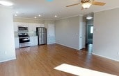 This single-floor home offers an open and airy layout that seamlessly connects the living and kitchen areas, creating a welcoming and functional space. The kitchen is outfitted with stainless steel appliances, including a range, microwave, and refrigerator, complemented by crisp white cabinetry and light stone countertops. The generous open living area flows naturally from the kitchen, making it well-suited for both everyday living and entertaining. Warm hardwood-style flooring runs throughout, adding a sense of cohesion and a touch of elegance to the interior. Crown molding lines the ceilings, lending a refined architectural detail, while recessed lighting and a classic ceiling fan with a light fixture ensure both comfort and ample illumination. Neutral gray walls provide a versatile backdrop that pairs well with a variety of décor styles. Interior doorways lead to additional rooms, hinting at a thoughtfully designed floor plan that balances open communal spaces with private quarters. Natural light filters into the space, enhancing the bright and clean aesthetic that defines this well-maintained home.