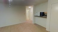 This single-floor residential unit offers a clean, functional layout well-suited for comfortable living. The main living area is finished with wall-to-wall carpeting in a neutral tone, creating a warm and inviting atmosphere. Smooth, light-colored walls throughout lend the space a bright, airy feel, complemented by ceiling-mounted lighting that ensures consistent illumination.

A defining feature of the layout is the open pass-through counter that connects the kitchen to the living area, providing a convenient breakfast bar or serving surface. The kitchen is equipped with a refrigerator and features upper cabinetry, offering practical storage solutions. The dark countertop provides a subtle contrast against the lighter surroundings.

A hallway visible from the main living area leads to additional rooms, hinting at a thoughtful floor plan that keeps living and private spaces well separated. A door at the end of the hallway — likely a bathroom or exterior entry — is framed by overhead lighting, adding depth to the overall layout.

The neutral color palette, consistent flooring, and straightforward design make this space highly versatile and easy to personalize. Overall, the unit presents a tidy, move-in-ready environment with a practical and efficient use of space throughout.
