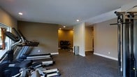 This single-floor fitness facility is designed to accommodate a variety of workout routines within a thoughtfully laid-out space. The area is divided into distinct zones, allowing for both cardio and strength training activities. Dark, durable carpet flooring covers the entire space, providing both comfort and practicality for exercise use. The walls are painted in warm taupe and neutral tones, creating an inviting yet focused atmosphere conducive to physical activity. Recessed lighting is installed throughout the ceiling, delivering consistent, even illumination across all workout areas. The main area houses cardio equipment, including rowing machines and step trainers, positioned conveniently near the entrance. A secondary alcove, warmly lit and partially separated by a structural column, contains a multi-tiered dumbbell rack stocked with free weights alongside a weight bench, forming a dedicated strength training zone. A hallway extends further into the space, suggesting additional rooms or utility areas beyond. White baseboards provide a clean contrast against the darker walls and flooring, contributing to a polished, well-maintained appearance throughout the facility.