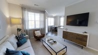 This 2-bedroom, 2-bathroom residence offers a thoughtfully designed single-level layout that blends comfort and style. The open-concept living and dining area serves as the heart of the home, bathed in natural light streaming through large windows dressed with elegant sheer curtains and plantation shutters. Crown molding adds a refined architectural detail throughout the space, complementing the neutral color palette and clean lines.

The living area features plush carpeting, a sleek glass-top coffee table, and a wall-mounted flat-screen television positioned above a rustic wood console, creating a warm and inviting focal point. A leather accent chair adds a touch of sophistication alongside the comfortable sofa adorned with teal and navy throw pillows.

The dining area flows seamlessly from the living space, illuminated by a contemporary chandelier and surrounded by windows that enhance the airy ambiance. The open layout between the kitchen and dining area creates an ideal setting for entertaining. The overall design strikes a balance between modern aesthetics and cozy livability, making this residence a welcoming and functional home.