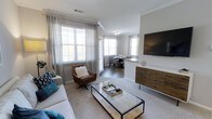 This 2-bedroom, 2-bathroom residence offers a thoughtfully designed single-level layout that blends comfort and style. The open-concept living and dining area serves as the heart of the home, bathed in natural light streaming through large windows dressed with elegant sheer curtains and plantation shutters. Crown molding adds a refined architectural detail throughout the space, complementing the neutral color palette and clean lines.

The living area features plush carpeting, a sleek glass-top coffee table, and a wall-mounted flat-screen television positioned above a rustic wood console, creating a warm and inviting focal point. A leather accent chair adds a touch of sophistication alongside the comfortable sofa adorned with teal and navy throw pillows.

The dining area flows seamlessly from the living space, illuminated by a contemporary chandelier and surrounded by windows that enhance the airy ambiance. The open layout between the kitchen and dining area creates an ideal setting for entertaining. The overall design strikes a balance between modern aesthetics and cozy livability, making this residence a welcoming and functional home.