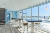 Located along Harbor Walk Avenue in Norfolk, Virginia, this single-floor coastal-inspired community space offers a bright and welcoming atmosphere throughout. The open-concept layout is unified by warm wood-look tile flooring, crisp white crown molding, and soft sky-blue accent walls that evoke a seaside aesthetic at every turn.

The kitchen is a standout feature, outfitted with dark shaker-style cabinetry, granite countertops, and stainless steel appliances including a full-size refrigerator. A large central island with seating for multiple guests serves as an ideal gathering spot, while shiplap wall detailing and an eye-catching cluster of Edison bulb pendant lights add a stylish, nautical-industrial touch. Open wood shelving completes the functional yet decorative design.

The adjoining lounge area is generously proportioned, furnished with navy blue wicker chairs and patterned cushions that reinforce the coastal theme. Large-scale nautical artwork adorns the walls, and recessed lighting keeps the space well-illuminated. A wall-mounted flat-screen TV and a shuffleboard table offer entertainment options for residents and guests alike.

Expansive floor-to-ceiling windows and French doors open to an outdoor balcony overlooking what appears to be a marina, flooding the interior with natural light and offering picturesque waterfront views. This space combines comfort, style, and function in a cohesive coastal design.