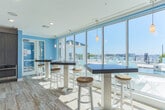 Located along Harbor Walk Avenue in Norfolk, Virginia, this single-floor coastal-inspired community space offers a bright and welcoming atmosphere throughout. The open-concept layout is unified by warm wood-look tile flooring, crisp white crown molding, and soft sky-blue accent walls that evoke a seaside aesthetic at every turn.

The kitchen is a standout feature, outfitted with dark shaker-style cabinetry, granite countertops, and stainless steel appliances including a full-size refrigerator. A large central island with seating for multiple guests serves as an ideal gathering spot, while shiplap wall detailing and an eye-catching cluster of Edison bulb pendant lights add a stylish, nautical-industrial touch. Open wood shelving completes the functional yet decorative design.

The adjoining lounge area is generously proportioned, furnished with navy blue wicker chairs and patterned cushions that reinforce the coastal theme. Large-scale nautical artwork adorns the walls, and recessed lighting keeps the space well-illuminated. A wall-mounted flat-screen TV and a shuffleboard table offer entertainment options for residents and guests alike.

Expansive floor-to-ceiling windows and French doors open to an outdoor balcony overlooking what appears to be a marina, flooding the interior with natural light and offering picturesque waterfront views. This space combines comfort, style, and function in a cohesive coastal design.