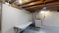 This unfinished basement presents a spacious, open-plan layout with significant potential for customization or buildout. The ceiling features fully exposed wooden floor joists and visible ductwork, along with galvanized HVAC ducts and electrical conduit running overhead, giving the space a raw, utilitarian character. The concrete floors are painted gray throughout, providing a clean and durable base across the entire area. The perimeter walls are constructed of painted concrete masonry, with evidence of water sealing treatments applied in several areas. A dedicated laundry zone is equipped with a standalone dryer and includes a long folding table, making it functional for everyday use. A separate enclosed room with its own door offers additional private storage or utility space. Small ground-level windows allow modest natural light to filter in, supplemented by bare-bulb pendant lighting and fluorescent fixtures. A utility area along one wall houses visible plumbing and mechanical components. The layout of the basement, as seen in the overhead model view, reveals an L-shaped floor plan with distinct zones. The space is well-suited for conversion into additional living areas, a workshop, or expanded storage, with all major systems clearly accessible for inspection or modification.