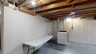 This unfinished basement presents a spacious, open-plan layout with significant potential for customization or buildout. The ceiling features fully exposed wooden floor joists and visible ductwork, along with galvanized HVAC ducts and electrical conduit running overhead, giving the space a raw, utilitarian character. The concrete floors are painted gray throughout, providing a clean and durable base across the entire area. The perimeter walls are constructed of painted concrete masonry, with evidence of water sealing treatments applied in several areas. A dedicated laundry zone is equipped with a standalone dryer and includes a long folding table, making it functional for everyday use. A separate enclosed room with its own door offers additional private storage or utility space. Small ground-level windows allow modest natural light to filter in, supplemented by bare-bulb pendant lighting and fluorescent fixtures. A utility area along one wall houses visible plumbing and mechanical components. The layout of the basement, as seen in the overhead model view, reveals an L-shaped floor plan with distinct zones. The space is well-suited for conversion into additional living areas, a workshop, or expanded storage, with all major systems clearly accessible for inspection or modification.