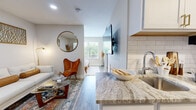 This thoughtfully designed 1-bedroom, 1-bathroom unit offers an efficient single-floor layout that maximizes every square foot. Light hardwood-style flooring flows throughout, anchoring a neutral, modern aesthetic. The open-plan living area is bright and welcoming, featuring recessed lighting and a window that brings in natural light. The kitchen runs galley-style with white shaker cabinets accented by gold hardware, subway tile backsplash, stainless steel appliances including a gas range and full-size refrigerator, and granite-look countertops that extend to a breakfast bar with barstool seating. The living room connects seamlessly to the kitchen, offering a comfortable lounge area with a wall-mounted TV. Two closets provide practical storage within the unit. The bedroom is a calm, minimal retreat with a large window overlooking greenery, providing abundant natural light and outdoor views. Warm-toned brass bedside lighting and clean white walls contribute to the serene atmosphere. The bathroom is finished with a white vanity featuring marble-look countertops, tile flooring, and a bright overhead light above a wide mirror, completing the clean, modern design of this well-appointed unit.