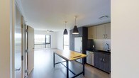 This single-floor space showcases a thoughtfully designed open-concept layout that seamlessly integrates the kitchen and living areas. The kitchen is outfitted with stainless steel appliances, including a refrigerator and dishwasher, complemented by two-tone cabinetry featuring dark lower units and light upper cabinets. A standout centerpiece is the butcher block island table with sleek black metal legs, ideal for dining or food preparation. Two matte black pendant lights hang above, adding a contemporary industrial touch. The dark tile backsplash provides a striking contrast against the lighter cabinetry. The open living area benefits from large floor-to-ceiling windows that flood the space with natural light and offer views of the surrounding landscape. A ceiling fan is installed in the living area, enhancing comfort. Light-toned flooring runs throughout, contributing to a bright and cohesive aesthetic. The overall ambiance is modern and minimalist, with a clean, neutral palette punctuated by thoughtful design details.