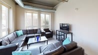 This single-floor loft-style unit blends industrial design with modern comfort throughout its well-organized layout. Exposed metallic ductwork runs along the ceilings, lending a distinctive urban character to the living spaces. The open-concept kitchen is outfitted with rich dark cabinetry, granite countertops, and a full suite of stainless steel appliances including a side-by-side refrigerator with ice and water dispenser, a smooth-top electric range, dishwasher, and microwave. Track lighting and a sliding glass door to an outdoor balcony add both functionality and natural light to the kitchen area. The spacious living room accommodates generous seating and is brightened by large windows overlooking the surrounding streetscape, creating an inviting gathering space. Multiple bedrooms offer flexible sleeping arrangements, with one room featuring bunk-style accommodation and a street-facing window, while another provides twin beds and wall art in a calm, neutral setting. A well-appointed bathroom includes a double vanity with granite countertops and dark cabinetry, complementing the kitchen's aesthetic. A dedicated in-unit laundry closet with a full-size washer and dryer adds everyday convenience. A hallway with polished concrete-style flooring connects the rooms seamlessly throughout the unit.
