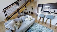 This thoughtfully designed two-floor space showcases a seamless open-concept layout that connects the living, dining, and kitchen areas into one cohesive and airy environment. Light hardwood flooring runs throughout the main level, complementing the warm tones of the natural wood staircase, which is accented by bold black metal hardware and wire-panel railings — a standout architectural feature that blends industrial and contemporary styles.

The living area offers a comfortable and welcoming atmosphere, centered around a light gray sofa accented with colorful throw pillows and anchored by a textured area rug. Just beyond, a compact dining nook provides an intimate space for meals, set against a backdrop of neutral walls adorned with tasteful artwork.

The kitchen is sleek and modern, featuring dark upper cabinetry, stainless steel appliances, and a white waterfall-edge island with bar seating — ideal for casual dining or entertaining. The layout maximizes functionality while maintaining a clean, contemporary aesthetic.

The second floor, accessible via the eye-catching staircase, expands the usable living space and adds a sense of verticality and openness to the overall design. Throughout the space, the interplay of light wood, dark metal accents, and neutral tones creates a modern yet inviting ambiance.
