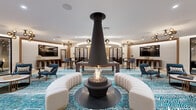 This beautifully designed amenity space delivers a sophisticated, resort-inspired experience across a single floor. At the heart of the main lounge, a striking suspended cone fireplace serves as the dramatic focal point, encircled by curved cream sectional seating and vibrant teal zebra-print carpeting that flows throughout. Gold globe pendant chandeliers cast a warm, ambient glow over an eclectic mix of mid-century modern furnishings, including sculptural lounge chairs, marble-topped coffee tables, and a full billiards table. A curved built-in bar area with counter seating adds a social hub, while a separate intimate alcove features a generous semi-circular gray sofa ideal for relaxed gatherings. The space also includes a dedicated golf simulator room, complete with a high-definition projection screen, synthetic turf flooring, and club storage — offering an engaging recreational retreat. Private conference and card rooms enclosed in sleek black-framed glass partitions provide quieter, functional settings for meetings or focused use. A formal dining area with rich wood furnishings and statement pendant lighting rounds out the offering. Curated black-and-white architectural photography adorns the walls, adding a gallery-quality aesthetic that ties together this dynamic, multi-functional amenity floor.