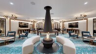 This beautifully designed amenity space delivers a sophisticated, resort-inspired experience across a single floor. At the heart of the main lounge, a striking suspended cone fireplace serves as the dramatic focal point, encircled by curved cream sectional seating and vibrant teal zebra-print carpeting that flows throughout. Gold globe pendant chandeliers cast a warm, ambient glow over an eclectic mix of mid-century modern furnishings, including sculptural lounge chairs, marble-topped coffee tables, and a full billiards table. A curved built-in bar area with counter seating adds a social hub, while a separate intimate alcove features a generous semi-circular gray sofa ideal for relaxed gatherings. The space also includes a dedicated golf simulator room, complete with a high-definition projection screen, synthetic turf flooring, and club storage — offering an engaging recreational retreat. Private conference and card rooms enclosed in sleek black-framed glass partitions provide quieter, functional settings for meetings or focused use. A formal dining area with rich wood furnishings and statement pendant lighting rounds out the offering. Curated black-and-white architectural photography adorns the walls, adding a gallery-quality aesthetic that ties together this dynamic, multi-functional amenity floor.