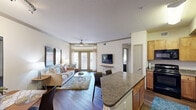 This efficiently designed single-story, one-bedroom, one-bathroom residence offers a thoughtful open-concept layout that maximizes its comfortable footprint. Rich hardwood-style flooring flows throughout the main living areas, anchoring the combined living, dining, and kitchen space in a warm, cohesive aesthetic.

The kitchen is well-equipped with warm-toned wood cabinetry, granite-style countertops, black appliances including a range, built-in microwave, dishwasher, and refrigerator. A peninsula island with bar seating provides additional prep space and serves as a natural divider between the kitchen and dining area.

The living room features a ceiling fan with light fixture and large French doors that invite abundant natural light while providing direct outdoor access. Crown molding adds a refined architectural detail throughout the main living space.

The bedroom is carpeted for comfort, features a tray ceiling, a ceiling fan, and large windows that bring in generous natural light. It connects directly to the full bathroom, which includes a single-sink vanity, a shower-tub combination, and hardwood-style flooring. A dedicated closet provides organized storage. A hallway connects the private areas of the residence to the main living space, completing this practical and comfortable floor plan.