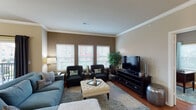 This single-story residence presents a thoughtfully designed layout that balances comfort and functionality across its key living spaces. Crown molding is a consistent architectural detail found throughout, lending a refined finish to each room.

The living room serves as a central gathering space, featuring hardwood floors, warm taupe walls, and a bank of three windows fitted with horizontal blinds that invite ample natural light. Dark-toned accent chairs and a tufted ottoman complement the soft blue sofa, creating an inviting atmosphere.

Adjacent to the living room, the dining area is anchored by a dark wood table beneath a chandelier, with a sliding glass door offering direct access to a balcony and outdoor views. The kitchen is well-equipped with rich cherry cabinetry, granite countertops, stainless steel appliances, a kitchen island with bar seating, and track lighting overhead.

The study provides a dedicated workspace with a large desk, accent chair, and a window with patterned drapery. The bedroom features plush carpeting, dual windows with outdoor views, and warm neutral tones. The bathroom is finished with granite countertops, a rich wood vanity, an oversized mirror, and a full shower-tub combination.