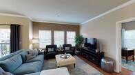 This single-story residence presents a thoughtfully designed layout that balances comfort and functionality across its key living spaces. Crown molding is a consistent architectural detail found throughout, lending a refined finish to each room.

The living room serves as a central gathering space, featuring hardwood floors, warm taupe walls, and a bank of three windows fitted with horizontal blinds that invite ample natural light. Dark-toned accent chairs and a tufted ottoman complement the soft blue sofa, creating an inviting atmosphere.

Adjacent to the living room, the dining area is anchored by a dark wood table beneath a chandelier, with a sliding glass door offering direct access to a balcony and outdoor views. The kitchen is well-equipped with rich cherry cabinetry, granite countertops, stainless steel appliances, a kitchen island with bar seating, and track lighting overhead.

The study provides a dedicated workspace with a large desk, accent chair, and a window with patterned drapery. The bedroom features plush carpeting, dual windows with outdoor views, and warm neutral tones. The bathroom is finished with granite countertops, a rich wood vanity, an oversized mirror, and a full shower-tub combination.