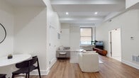 This single-floor unit presents a thoughtfully designed interior with a consistent modern aesthetic throughout. The living area serves as the heart of the space, featuring exposed ceiling beams, recessed lighting, and warm hardwood flooring that extends throughout the unit. A window allows natural light to filter in, illuminating the neutral, cream-toned walls and creating an inviting atmosphere. The seating area accommodates comfortable furnishings alongside a mid-century style media console.

The kitchen is well-appointed with white shaker cabinetry extending to the ceiling, clean light countertops, and a full suite of stainless steel appliances including a refrigerator, range, microwave, and dishwasher. A single-basin sink with a contemporary faucet anchors the workspace.

Two bedrooms offer restful retreats, each finished with the same hardwood flooring and bright white walls. One bedroom includes dual closet doors, providing ample storage, while both rooms feature understated, warm lighting.

The bathroom is distinguished by classic white hexagonal floor tiles and a wall-mounted sink paired with a round mirror, lending a clean, timeless character. The overall ambiance of the unit is minimal, bright, and cohesive.