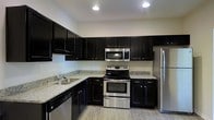 Located at 1821 Buckeye Drive in Richmond Hill, Georgia, this single-story home offers a clean, modern aesthetic throughout its layout. The kitchen serves as a standout feature of the space, showcasing rich, dark espresso-toned cabinetry that extends from base to upper wall units, providing generous storage capacity. Light granite countertops offer a striking contrast against the dark cabinetry, creating a bold and contemporary visual appeal. The kitchen is outfitted with a full suite of stainless steel appliances, including a French door refrigerator, electric range with oven, over-the-range microwave, and dishwasher. An L-shaped counter layout maximizes workspace efficiency, while a double-basin sink is positioned beneath the cabinetry for convenience. A pendant light fixture hangs gracefully over the kitchen area, adding a warm, inviting glow that complements the recessed lighting throughout. The light wood-tone flooring runs seamlessly through the space, lending a sense of continuity and warmth. Neutral wall tones keep the interior bright and versatile, making the space well-suited for a variety of furnishing styles. The overall design balances function and style in a well-appointed, move-in ready setting.