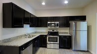 Located at 1821 Buckeye Drive in Richmond Hill, Georgia, this single-story home offers a clean, modern aesthetic throughout its layout. The kitchen serves as a standout feature of the space, showcasing rich, dark espresso-toned cabinetry that extends from base to upper wall units, providing generous storage capacity. Light granite countertops offer a striking contrast against the dark cabinetry, creating a bold and contemporary visual appeal. The kitchen is outfitted with a full suite of stainless steel appliances, including a French door refrigerator, electric range with oven, over-the-range microwave, and dishwasher. An L-shaped counter layout maximizes workspace efficiency, while a double-basin sink is positioned beneath the cabinetry for convenience. A pendant light fixture hangs gracefully over the kitchen area, adding a warm, inviting glow that complements the recessed lighting throughout. The light wood-tone flooring runs seamlessly through the space, lending a sense of continuity and warmth. Neutral wall tones keep the interior bright and versatile, making the space well-suited for a variety of furnishing styles. The overall design balances function and style in a well-appointed, move-in ready setting.