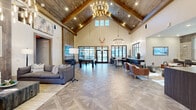 Located at 2116 Pleasant Road in Fort Mill, SC, this single-floor community clubhouse offers a generously sized, open-concept interior designed with a refined rustic-modern aesthetic. The centerpiece of the space is a dramatic great room with soaring cathedral ceilings clad in warm wood paneling and anchored by exposed timber beams, all illuminated by statement chandelier lighting. Geometric-patterned tile flooring runs throughout, adding visual depth and sophistication. Large black-framed glass doors and transom windows flood the interior with abundant natural light while offering views of the surrounding outdoor greenery. The main lounge area features a mix of plush gray sofas and leather accent chairs arranged around a central gathering space, with a pool table providing a focal point for recreation. A dedicated lounge nook offers additional seating with a wall-mounted display, opening into a secondary room with a game table and ottomans. The kitchen-style counter area includes a coffee station, dark cabinetry with herringbone tile backsplash, and bar-height seating — ideal for casual interaction. A private office is also included within the layout. The welcoming entry foyer features high ceilings, a circular chandelier, and a large art piece, setting an impressive tone upon arrival.