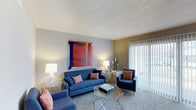 This single-floor residence offers a comfortable and thoughtfully designed living space with a clean, modern aesthetic. The living room serves as the focal point of the home, showcasing plush carpeted flooring that adds warmth underfoot. A bold, vibrant abstract artwork anchors one of the neutral-toned walls, adding a striking pop of color and artistic character to the room. The space is furnished with a deep navy blue sofa and coordinating armchairs, accented with patterned throw pillows in warm red and orange tones, creating a welcoming and cohesive color palette. A sleek glass coffee table and accent side tables with chrome-finished legs lend a contemporary touch to the décor. Floor-to-ceiling vertical blinds frame a large sliding glass door, allowing generous natural light to flood the interior while offering a glimpse of the greenery beyond. The textured ceiling adds subtle dimension overhead, while soft ambient lighting from stylish table lamps ensures a warm and inviting atmosphere throughout the day and evening. The overall ambiance of this residence is bright, airy, and move-in ready, blending comfort with modern style in a cohesive single-level layout.
