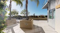 Located on Chandler Boulevard in the heart of Los Angeles, this rooftop terrace offers an expansive outdoor retreat with a distinctly Southern California character. The level is finished with large-format light-toned tiles that reflect the abundant natural sunlight, creating a bright and open atmosphere throughout the space. Tall palm trees rise from substantial concrete planters anchored across the terrace, providing organic vertical interest and shade. Lush greenery, including dense climbing shrubs along the building's perimeter walls, adds a layer of vibrant landscaping that softens the clean, modern architectural lines. A dedicated seating area, outfitted with lounge chairs and shaded by large patio umbrellas, offers a comfortable setting for relaxation while taking in the panoramic views of the surrounding hillsides and Los Angeles skyline. The terrace is bordered by low perimeter walls that frame unobstructed sightlines to the mountains beyond. The adjacent building facade features floor-to-ceiling glazing with aluminum framing and an upper-level glass-railed balcony, reflecting the contemporary design aesthetic of the overall structure. Walkways lined with built-in planters guide movement across the terrace, connecting covered corridor access points to the open-air gathering areas. This rooftop space combines thoughtful landscaping with modern design to deliver an inviting outdoor environment.
