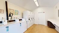 Located at 1100 Fairhaven Ave in Santa Ana, California, this communal laundry facility is a thoughtfully designed, single-room space that prioritizes functionality and comfort. The room features a vaulted ceiling with crown molding, giving it an airy, open feel that belies its compact footprint. Light wood-look flooring runs throughout, contributing to a clean and welcoming atmosphere.

The laundry area is equipped with multiple top-load washing machines and a bank of stacked dryer units lining one wall, providing ample capacity for residents. A standout feature is the generous wall-mounted folding counter finished in a marble-style surface, offering a convenient and attractive workspace for sorting and folding laundry. A corkboard bulletin board is mounted nearby, serving as a communication hub for residents.

Natural light pours in through a set of white French doors that open to a lush outdoor courtyard, enhancing the brightness and ventilation of the space. Overhead, fluorescent lighting ensures consistent illumination throughout the day and evening. The room also includes a small utility table and a fire safety station, reflecting attention to both practical use and resident safety. The overall ambiance is tidy, functional, and well-maintained, making this laundry room a valuable and pleasant shared amenity.