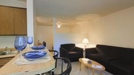 This single-floor space is thoughtfully arranged with an open-plan design that integrates the dining and living areas into one cohesive environment. The dining area is positioned toward the front, featuring a round light-wood table with curved black chairs and place settings that add a welcoming touch. The living area flows naturally from the dining space and is furnished with dark, plush armchairs and a sofa arranged around a wooden oval coffee table, creating a relaxed and inviting atmosphere. A warm floor lamp provides soft ambient lighting, complementing the neutral tones of the walls and carpeted flooring throughout. The ceiling features a textured finish and includes a ceiling fan, which adds both a functional and casual element to the space. A partial wall division subtly separates the two zones while maintaining an open, airy feel across the layout. The overall ambiance is understated and practical, with a neutral color palette of beige and white walls that allows the dark furnishings and blue accent pieces to stand out. The space is well-suited for everyday living, offering a comfortable blend of dining and lounging areas within a compact and efficiently designed floor plan.