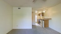This single-floor residence offers a straightforward and functional layout designed for comfortable everyday living. The living area features wall-to-wall carpeting that adds warmth underfoot, with a ceiling fan overhead to ensure year-round comfort. A textured ceiling runs throughout, adding a classic touch to the interior. The space opens naturally toward the kitchen, which is positioned at the heart of the home. The kitchen is well-appointed with wood-toned cabinetry, a range, a refrigerator, and in-unit washer, providing added convenience. A peninsula-style breakfast bar with a laminate countertop serves as both a workspace and a casual dining spot, creating a seamless transition between the kitchen and the main living area. A second ceiling fan in the kitchen keeps the area well-ventilated. Natural light filters in through a window near the kitchen, brightening the space. The neutral color palette of cream and beige tones throughout creates a clean, cohesive atmosphere that is easy to personalize. The overall ambiance is practical and inviting, making it a comfortable and move-in ready space.