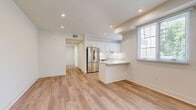 This well-appointed single-floor unit offers a clean, modern aesthetic throughout, highlighted by light hardwood flooring, crisp white walls, and recessed lighting that create a bright and welcoming atmosphere. The open-concept living and kitchen area forms the heart of the unit, where large windows fitted with white horizontal blinds allow abundant natural light and views of surrounding greenery. The kitchen is thoughtfully designed with white shaker-style cabinetry, white subway tile backsplash, light quartz countertops, a peninsula with bar seating, and a full suite of stainless steel appliances including a refrigerator, electric range, and dishwasher. The spacious living area flows seamlessly from the kitchen, offering ample room for flexible furniture arrangements. The bedrooms are well-sized, featuring light hardwood or plush carpeted flooring, built-in sliding-door closets with shelving, and windows that provide natural light. The full bathroom stands out with eye-catching large-format hexagonal floor tile in a classic cream-and-dark-border pattern, complemented by a white vanity, a mirror with overhead lighting, and a full tub-shower combination. A central hallway connects all areas of the unit efficiently, completing this thoughtfully designed living space.
