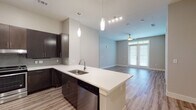 This single-floor residence features a contemporary open-concept design that seamlessly connects the kitchen and living area. The kitchen is well-appointed with dark espresso cabinetry, white quartz countertops, a tile backsplash, and a full suite of stainless steel appliances, including a refrigerator with ice maker, range, and dishwasher. A large central island with an undermount sink and pendant lighting above serves as both a functional workspace and a natural gathering point. The living area extends from the kitchen and benefits from natural light through sliding glass doors, complemented by a ceiling fan overhead. Wood-look plank flooring runs throughout the main living spaces, adding warmth and continuity. The bedrooms are carpeted and receive ample natural light through multiple windows with blinds. The primary bathroom includes dark vanity cabinetry with white countertops, tile flooring, and access to a walk-in closet with built-in shelving. A secondary bathroom offers a similarly clean, minimalist design with matching tile floors and dark cabinetry. The overall aesthetic is modern and cohesive, with neutral tones and clean lines throughout.