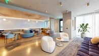Located at 320 W South St in downtown Raleigh, NC, this thoughtfully designed single-floor amenity space offers a generous footprint across a living room, game room, hallway, and additional lounge area. The centerpiece of the space is a dramatic oval cove ceiling with warm ambient uplighting that anchors the main lounge, where a large sectional sofa, rounded olive-green velvet armchairs, and a curated mix of accent furniture create an inviting, sophisticated atmosphere. A sweeping curved counter with pendant lighting serves as a bar or service station, adorned with tasteful artwork and greenery. Adjacent to the main lounge, a dining and café area features sleek black furniture set against large windows overlooking lush tree canopies, bathing the space in natural light. A warm timber slatted partition adds organic texture while maintaining visual openness. The hallway is defined by a striking full-height marble feature wall and a brass-framed magazine display unit, leading to a series of glass-walled private rooms with gold-toned frames. Smaller lounge nooks throughout offer intimate seating with botanical art and warm-toned furnishings. Polished concrete flooring flows seamlessly throughout, tying together a refined, contemporary aesthetic that balances comfort with modern elegance.