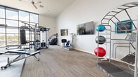 Located at 4333 N 6th Dr in Phoenix, AZ, this fitness center offers a thoughtfully designed workout space with impressive architectural details and a full complement of exercise equipment. The single-floor facility is characterized by soaring ceilings that create an open, airy atmosphere, while expansive floor-to-ceiling windows along one wall flood the space with abundant natural light and provide views of the surrounding neighborhood.

The interior features neutral-toned carpet flooring throughout and clean white walls that contribute to a bright, modern aesthetic. A ceiling fan provides additional air circulation, complementing the overall comfort of the space.

The fitness area is comprehensively equipped to support a wide variety of workout routines. Strength training options include a Synrgy360 functional training rig, a cable machine, a Smith machine with free weights, and an adjustable weight bench. A dedicated dumbbell rack and weight plates are also available. For flexibility and functional fitness, a tiered rack holds multiple stability and medicine balls in assorted sizes. Cardio equipment, including a step machine, rounds out the offerings. Wall-mounted flat-screen televisions are positioned throughout the space for entertainment during workouts. Full-length mirrors on one wall allow users to monitor their form, enhancing the overall training experience.