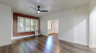 Located in the heart of Hollywood at 1775 N Orange Dr, Los Angeles, this well-appointed single-floor unit offers a thoughtfully designed layout comprising a living room, kitchen, hallway, and bathroom. The living room serves as the focal point of the space, showcasing a striking exposed brick accent wall framed by large multi-pane windows that invite lush greenery views and generous natural light. Rich, dark hardwood floors extend throughout, complemented by crisp white walls and a sleek black ceiling fan that adds a modern industrial touch. An elegant arched doorway connects the hallway, lending a classic architectural detail that adds character to the interior. The kitchen is efficiently laid out in a galley style, featuring white shaker cabinets with black hardware, dark granite countertops, a stainless steel sink with a matte black faucet, and full-size appliances — offering both style and functionality. The bathroom presents a clean, all-white aesthetic with classic penny tile flooring, a combination tub and shower, and matte black fixtures that tie the design together cohesively. Throughout the unit, consistent hardwood flooring and a neutral color palette create a unified, modern ambiance ideal for comfortable city living.