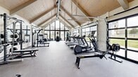 Located in Bozeman, Montana, this well-appointed fitness center offers a spacious, single-floor layout designed to support a comprehensive workout experience. The room's most striking architectural feature is its soaring vaulted ceiling finished with warm wood planking, which lends the space an open, airy quality and a polished aesthetic uncommon in typical fitness facilities.

Natural light floods the interior through multiple full-panel glass garage doors that line one side of the facility, framing views of the surrounding Montana landscape, including open green space and distant mountain scenery. These doors also allow for ventilation and create a seamless visual connection to the outdoors.

The fitness center is thoroughly equipped for both cardiovascular and strength training. The cardio section includes a row of treadmills, elliptical machines, a rowing ergometer, and a stationary spin bike. For strength training, the space features a power rack with a full barbell and plate set, an adjustable weight bench, a multi-function cable machine, and a well-stocked dumbbell rack. A large wall mirror on one end enhances the sense of space and allows for form checks during workouts.

Durable rubber-blend flooring covers the entire area, providing traction and cushioning. Recessed lighting supplements natural light throughout the day. The facility also includes a dedicated entry door, making it a self-contained, fully functional fitness center.
