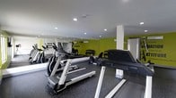 This fitness center is designed to deliver a focused and energizing workout environment. The space is outfitted with a solid selection of cardio equipment, including treadmills and elliptical machines from Spirit and other leading brands, providing users with versatile options for aerobic training. Additional exercise equipment, including stationary bikes and strength-training machines, rounds out the offerings for a comprehensive fitness experience.

The interior features bold lime-green accent walls adorned with motivational text, creating a vibrant and motivating atmosphere. Full-length mirrors line one wall, enhancing the sense of openness while allowing users to monitor their form during exercise. Recessed lighting in the white ceiling ensures the space is well-lit throughout. The flooring consists of durable rubber gym matting, offering both safety and comfort underfoot.

A structural column is positioned within the room, and a storage unit is visible along one wall. The layout is thoughtfully arranged to allow for comfortable movement between machines. Overall, this fitness center offers a clean, modern, and well-maintained environment suitable for a range of fitness routines and skill levels.