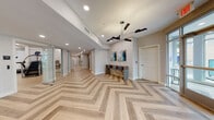 Located at 59 2nd Ave in Raritan, NJ, this single-floor amenity space combines modern design with high-end finishes to create a welcoming and functional environment. The main lounge area makes an immediate impression with its striking chevron-patterned hardwood flooring, a warm wood-paneled accent wall, and a sleek electric fireplace topped by a wall-mounted display screen. Large floor-to-ceiling windows flood the space with natural light and offer views of the surrounding streetscape. Seating is thoughtfully arranged with a cream sofa, mid-century modern accent chairs, and a chic bench, all anchored by a patterned area rug. A built-in kitchenette with wood cabinetry, a sink, coffee station, and compact refrigerator provides convenient amenities for residents. The corridors feature the same distinctive chevron hardwood flooring, sculptural black pendant lighting, curated wall art, and a built-in counter workspace with bar-height seating. The expansive fitness center is outfitted with a comprehensive range of equipment — including strength machines, treadmills, stair climbers, and cycling bikes — all beneath dramatic curved LED lighting. Textured accent walls, light wood flooring, and large windows keep the fitness area bright and energizing throughout.