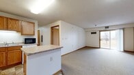 This single-floor unit offers a functional and straightforward layout across two bedrooms and two bathrooms. The combined kitchen and living area forms the heart of the space, where the kitchen is outfitted with warm oak cabinetry, a laminate countertop, white appliances including a refrigerator and range, a built-in microwave, and a central peninsula that provides additional counter space and separates the kitchen from the carpeted living area. Tile flooring anchors the kitchen zone, while neutral carpet extends throughout the living and bedroom areas. The living room receives natural light through a sliding glass door that opens to a private balcony with outdoor views of the surrounding neighborhood. Both bedrooms are modestly sized, carpeted, and finished with warm wood trim; the second bedroom features a large window that further enhances natural light. A hallway connects the bedrooms to the two bathrooms, each featuring oak vanities with solid-surface countertops and tile flooring. One bathroom includes a dedicated walk-in shower stall. Textured ceilings and warm wood door and window trim are consistent throughout, lending a cohesive and understated character to the overall space.