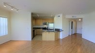 This single-floor space showcases a thoughtfully designed open-concept layout that seamlessly connects the kitchen and living areas. The kitchen is well-appointed with light maple cabinetry offering ample storage, dark granite countertops, and a full suite of stainless steel appliances, including a refrigerator, range, and built-in microwave. A curved breakfast bar extends from the kitchen, providing additional counter space and a casual dining option while serving as a natural transition between the kitchen and living areas. The kitchen features tile flooring, while the adjoining living space is finished with warm, light-toned hardwood floors that enhance the overall brightness of the room. Recessed lighting in the kitchen ensures even illumination throughout. Crown molding details add a polished touch to the space. A hallway visible from the main living area suggests additional rooms beyond, contributing to the functional flow of the layout. The overall ambiance is clean, modern, and practical, making it a versatile and welcoming living environment.