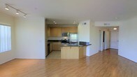 This single-floor space showcases a thoughtfully designed open-concept layout that seamlessly connects the kitchen and living areas. The kitchen is well-appointed with light maple cabinetry offering ample storage, dark granite countertops, and a full suite of stainless steel appliances, including a refrigerator, range, and built-in microwave. A curved breakfast bar extends from the kitchen, providing additional counter space and a casual dining option while serving as a natural transition between the kitchen and living areas. The kitchen features tile flooring, while the adjoining living space is finished with warm, light-toned hardwood floors that enhance the overall brightness of the room. Recessed lighting in the kitchen ensures even illumination throughout. Crown molding details add a polished touch to the space. A hallway visible from the main living area suggests additional rooms beyond, contributing to the functional flow of the layout. The overall ambiance is clean, modern, and practical, making it a versatile and welcoming living environment.