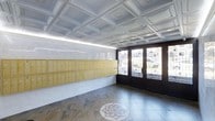 This building lobby combines classic architectural details with functional design, creating a welcoming and refined entry experience. The ceiling is a standout feature, showcasing an ornate coffered grid pattern finished in soft white and pale blue tones, accented by recessed lighting that evenly illuminates the space. Along one wall, a full bank of golden-yellow mailboxes provides a bold pop of color and serves the building's residents with a organized, practical layout. The flooring is composed of natural stone tiles in earthy, varied tones, and is highlighted by a striking circular mosaic medallion inlaid at the center of the floor — a testament to the craftsmanship and attention to detail incorporated into the space. The entry is framed by tall, dark-framed double doors fitted with frosted decorative glass panels featuring intricate scrollwork and floral ironwork details, allowing soft, diffused natural light to filter into the lobby. The walls are finished in a smooth, clean plaster, contributing to the airy and open atmosphere. Together, these elements create a lobby that feels both grand and inviting, reflecting a building with strong architectural character and thoughtful design.