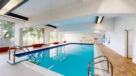Located at 190 Lees Ave in Ottawa, Ontario, this single-floor aquatic facility offers a well-appointed indoor swimming pool environment spanning a generous footprint. The centerpiece of the space is a large, clearly marked rectangular swimming pool with vivid blue water, white tile lining, and lane markings on the pool floor. The pool area features soaring vaulted ceilings with navy blue accent beams, creating a bright and airy atmosphere. Floor-to-ceiling windows along one side of the pool flood the space with natural light and provide views of the surrounding greenery outdoors. Safety equipment, including life rings and rescue poles, is mounted along the perimeter walls, and clear signage communicates pool rules throughout. Warm-toned terracotta tile flooring surrounds the pool deck, complemented by built-in bench seating. Connecting hallways lead to the changeroom and bathroom facilities, which feature the same terracotta tile flooring, private toilet stalls, open shower stalls, vanity with sink and mirror, and fluorescent overhead lighting. The overall design is functional and well-maintained, providing a complete aquatic amenity with all supporting facilities conveniently accessible within a single, thoughtfully organized floor plan.