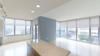 Located in Dallas, this contemporary single-floor space offers a thoughtfully designed open layout that maximizes both functionality and comfort. The interior is bathed in natural light, courtesy of expansive floor-to-ceiling windows wrapped around the main living area, fitted with neutral roller shades for adjustable privacy. These large windows frame views of the surrounding urban landscape, creating a seamless connection between the interior and the city outside.

A striking soft blue accent wall serves as a central architectural feature, adding depth and visual interest to the otherwise clean, minimalist palette. The crisp white ceilings are equipped with recessed lighting, providing even illumination throughout the space. Smooth, light-toned flooring runs continuously throughout, lending a sense of openness and cohesion.

The kitchen area is anchored by a generous granite-topped counter island with warm wood cabinetry beneath, offering ample workspace and storage. The granite surface features subtle speckled tones that complement the neutral color scheme of the space.

Overall, the space exudes a modern, polished ambiance with its combination of clean lines, high-quality finishes, and abundant natural light — making it a versatile and inviting environment well-suited for a variety of uses.